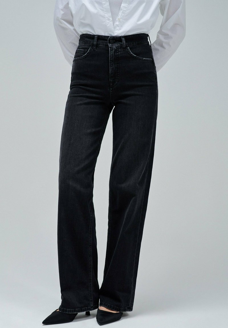 Persona que lleva unos pantalones rectos de talle alto de color negro, una camisa blanca abotonada metida por dentro y unos zapatos de tacón negro con punta puntiaguda, de pie contra un fondo gris.