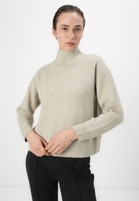 Maglione a coste di un beige chiaro, con collo alto, spalle scese e maniche lunghe. Indossato con pantaloni neri, che mostrano un tessuto testurizzato.