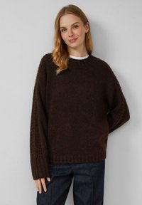 Donna con lunghi capelli biondi che indossa un maglione di lana marrone scuro e jeans scuri, in piedi contro uno sfondo grigio chiaro.