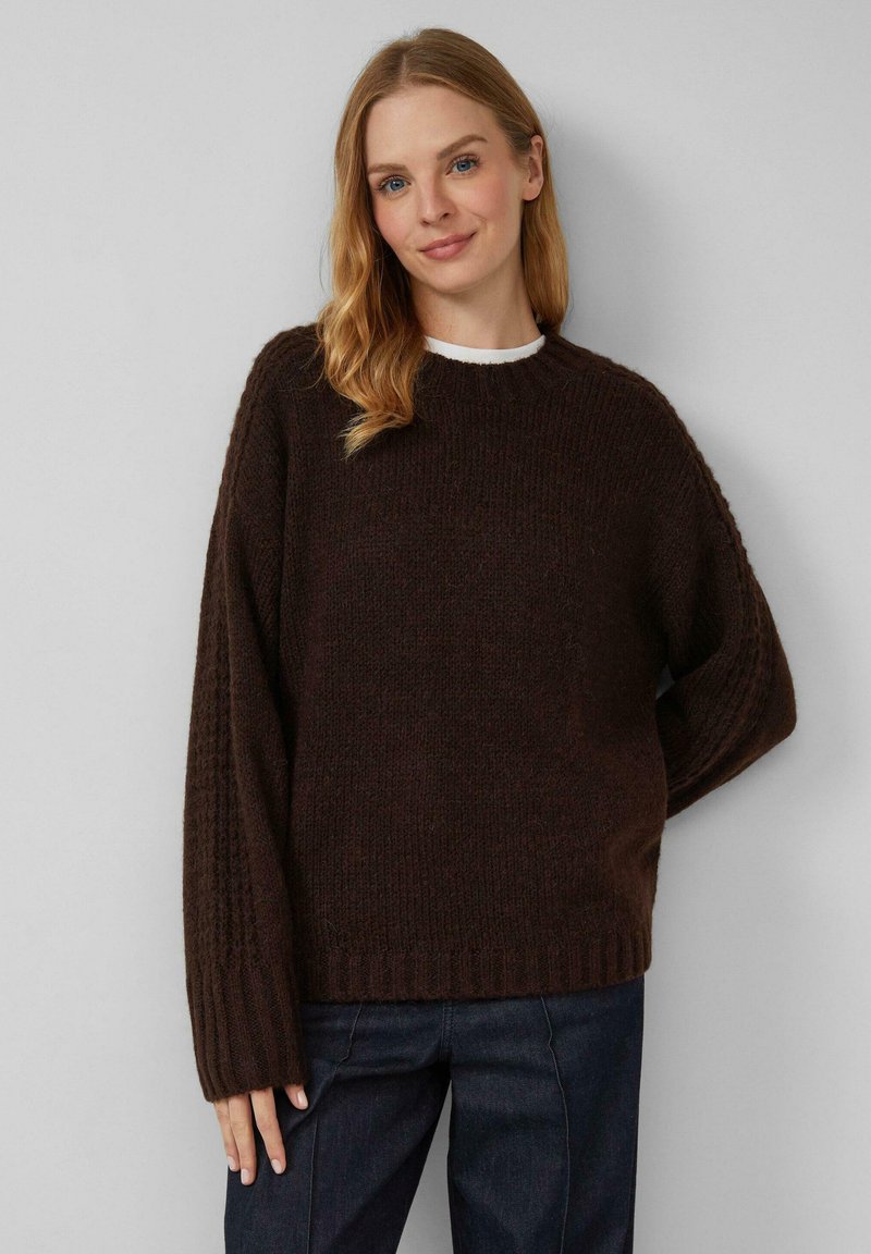 Donna con lunghi capelli biondi che indossa un maglione di lana marrone scuro e jeans scuri, in piedi contro uno sfondo grigio chiaro.