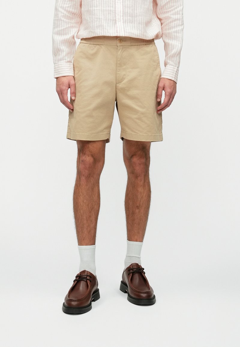 Shorts en coton beige avec une coupe décontractée, dotés de poches latérales et de bords retroussés. Associés à des chaussures en cuir marron et des chaussettes blanches.