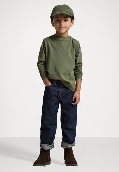 Zöld hosszúujjú póló logóval, sötétkék, felhajtott szárú farmernadrág és barna csizmák. A gyermek egy megfelelő zöld sapkát visel.