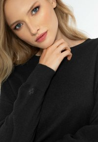 Schwarzer gestrickter Pullover mit Rundhalsausschnitt und langen Ärmeln. Besonders hervorzuheben ist ein kleines gesticktes Design am Bündchen.