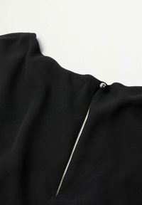 Tissu de vêtement noir avec un gros plan sur un petit bouton argenté fixant une ouverture fendue verticale étroite au niveau de l'encolure.