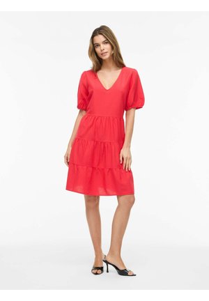 Femme portant une robe rouge longueur genoux avec des manches bouffantes et un décolleté en V, associée à des mules noires à talons ouverts, debout sur fond blanc.