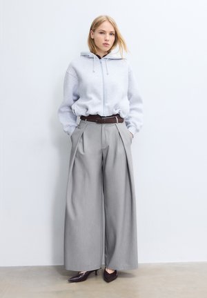Jeune femme portant un sweat à capuche gris clair zippé, un pantalon gris à jambes larges avec une ceinture marron, et des talons pointus foncés, debout contre un mur blanc.