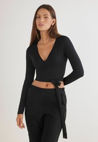 Černý top s dlouhými rukávy, véčkovým výstřihem a mašlí v pase. Materiál je hladký a lehký. Vyznačuje se vysokým pohodlím a je kombinován s černými kalhotami.