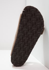 Brun sandal med en strukturerad svart gummisula, med ett vågmönster för grepp. Snedvy som visar det mjuka materialet och designen.