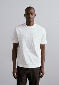 T-shirt blanc à manches courtes en mélange de coton avec un design de logo texturé sur le devant. Assorti avec un pantalon à rayures verticales foncées.