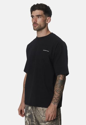 Man met kort donker haar en getatoeëerde arm draagt een zwart oversized t-shirt en camouflagebroeken, kijkend naar links.