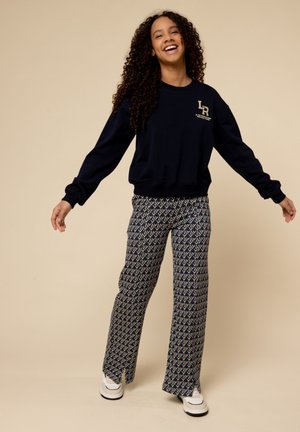 Marineblauwe sweatshirt met logo, gecombineerd met gestreepte flared broek met geometrische vormen in marineblauw, wit en roze. Witte sneakers maken de look af.