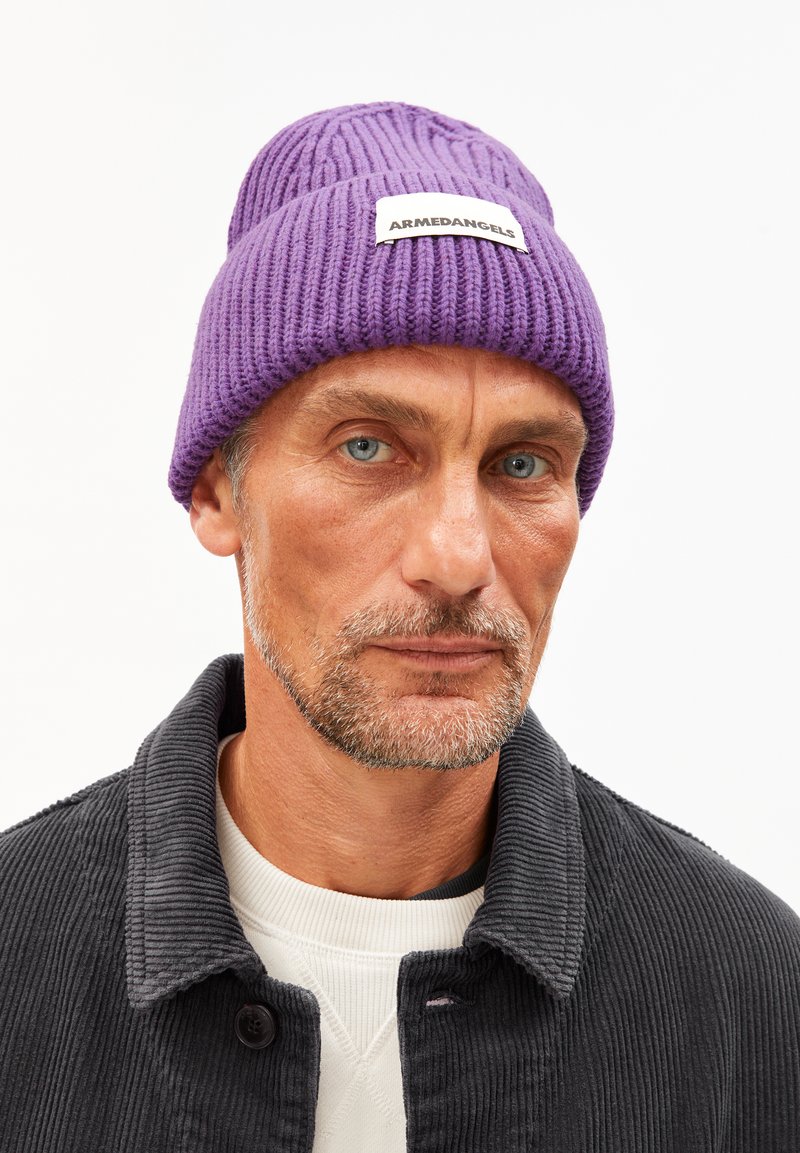 Mann mit leichtem Bart und blauen Augen, der eine lila Strickmütze mit dem Aufdruck "ARMEDANGELS", eine dunkle Cordjacke und ein weißes Rundhals-T-Shirt trägt.