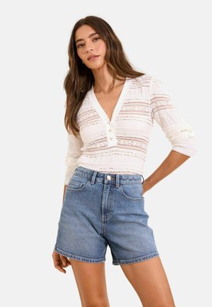 Naine, kes kannab valget tekstuuriga nööbitavat pikka varrukaga toppi ja kõrge vöökohaga siniseid teksapükse, seisab ühe käega puusal.