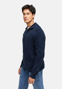 Marineblå zip-up trøje med ribbet tekstur, lange ærmer og ståkrave, sammen med lyseblå jeans.