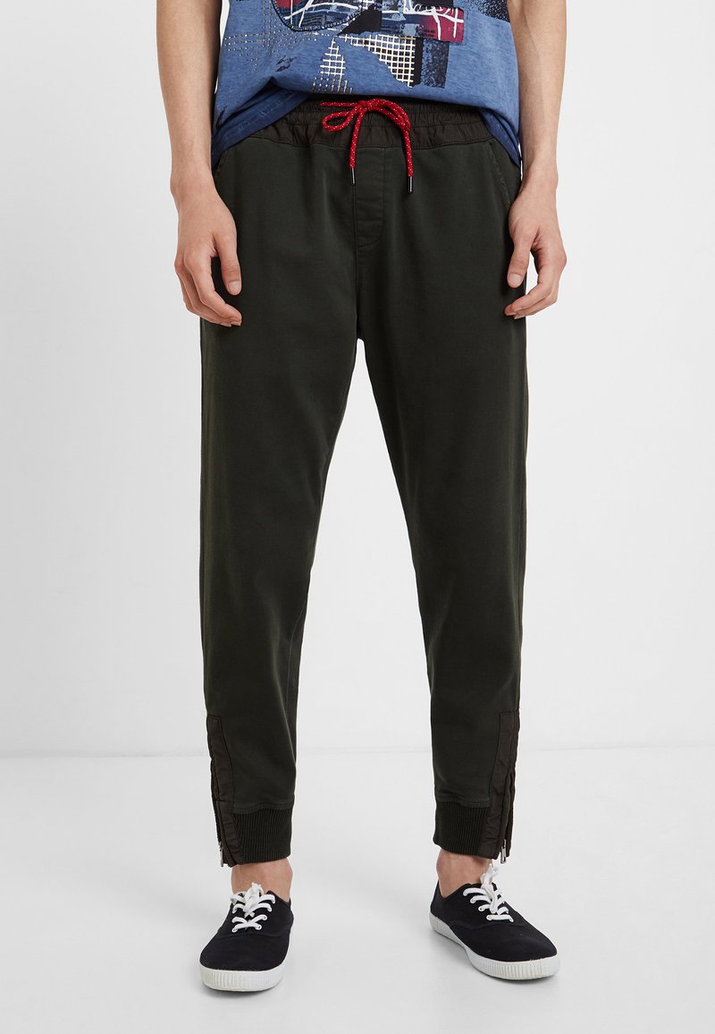 Dunkelgrüne Hose mit einem Kordelzugbund, roten Akzenten, tailliertem Design, seitlichen Reißverschlüssen und gerippten Knöchelabschlüssen. Kombiniert mit navyblauen Schuhen.