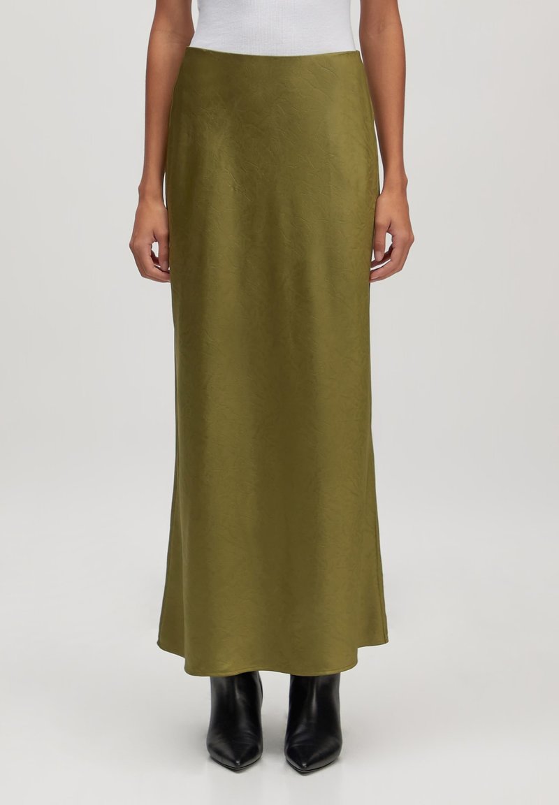 Olijfgroene maxi-rok van gladde stof, met een lange, vloeiende silhoette. Gekleed met spitse zwarte enkellaarsjes.
