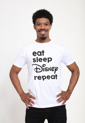 Uśmiechnięty mężczyzna z krótkimi kręconymi włosami stoi z rękami na biodrach, ubrany w białą koszulkę z czarnym napisem "eat sleep Disney repeat" na jednolitym tle.