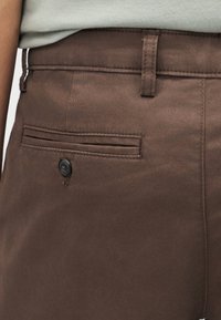 Καφέ παντελόνι με κουμπωμένη πίσω τσέπη που φοριέται με ανοιχτόχρωμο πουκάμισο, περασμένο μέσα.