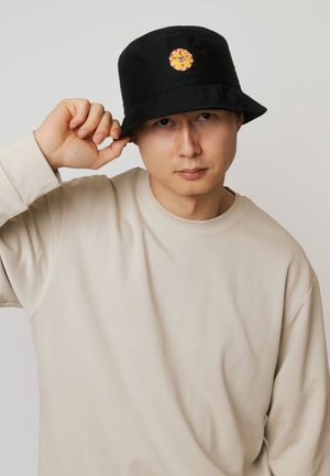 Mężczyzna w beżowej bluzie i czarnym kapeluszu typu bucket z haftowanym pomarańczowym słońcem, poprawiający rondo czapki na jasnym tle.