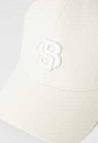 Bílá baseballová čepice s vyšitým logem "8". Má strukturovaný tvar, texturovanou látku a prošívané detaily podél kšiltu.
