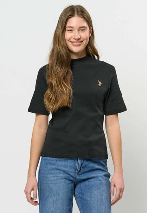 Ung kvinde med langt brunt hår iført en sort kortærmet skjorte med et lille broderet logo og blå jeans, smilende.