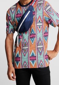 Camicia a maniche corte con un motivo geometrico colorato in arancione, teal e rosa. Abbinata a una borsa a tracolla blu navale con una cerniera.