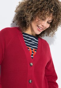 Roter Strickcardigan mit gerippter Textur, V-Ausschnitt und drei schwarzen Knöpfen. Über einem navyblauen gestreiften Shirt mit orangefarbener Schrift getragen.