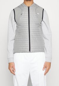 Grå vadderad väst med dragkedja fram, kontrasterad med grå ärmar. Har en logotyp på vänster bröst och en slät yta.