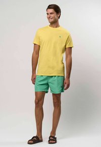Kollane puuvillane t-särk väikese rohelise logoga, koos piparmündiroheliste lühikeste pükste ja pruunide libisemisega sandaalidega. Lihtne disain, igapäevane lõige.