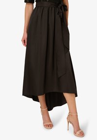 Czarna satynowa spódnica midi z plisami i związanym paskiem w talii. Posiada asymetryczny dół i noszona jest z nagimi, wiązanymi sandałami na obcasie.