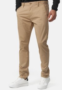 Chinos de algodón beige con corte recto, cierre de cremallera y botón, bolsillos laterales inclinados y detalles de costura mínimos. Llevados con zapatillas blancas.