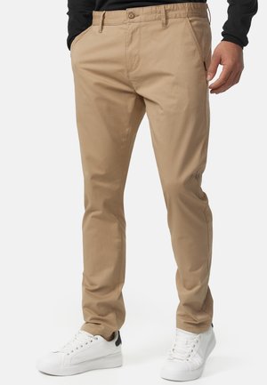 Beige cotton chinos with a straight-leg cut, zip and button closure, slanted side pockets, and minimal stitching details. Worn with white sneakers.