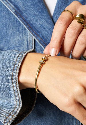 Bracciale rigido color oro con design liscio e arrotondato, caratterizzato da accenti decorativi e tre piccole pietre trasparenti. Indossato su un polso.