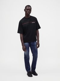 Homme noir debout portant un t-shirt noir Dsquared2, un jean bleu délavé et des baskets noires sur un fond blanc uni.