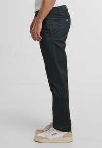 Schwarze gerade geschnittene Hose aus Baumwollmischgewebe, ausgestattet mit Seitentaschen und einem subtilen strukturierten Muster. Getragen mit weißen und beigen Sneakers.