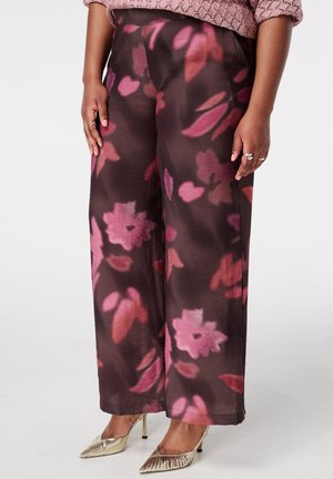 Pantalon à jambes larges avec des motifs floraux roses et rouges sur un fond sombre, accompagné de chaussures à talons hauts dorées et de bagues aux deux mains.
