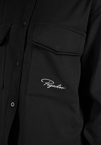 Camicia nera con due tasche sul petto, chiusura con bottoni e logo "Pegador" ricamato sulla tasca sinistra. Tessuto liscio, design classico.