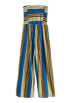 Ung kvinde med langt mørkt hår iført en stropløs, stribet jumpsuit i blå, gul, brun og cremefarvede nuancer, stående foran en ensfarvet væg.