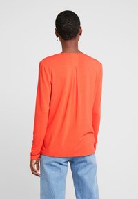 Camiseta naranja de manga larga con escote redondeado, detalle de pliegue en la parte trasera y corte ligeramente holgado, combinada con jeans de color azul claro.