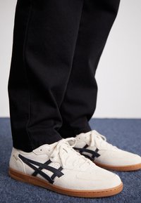 Ljus krämfärgade mockasneakers med mörkblå detaljer, med en strukturerad gummisula och klassisk snörning, i kombination med svarta byxor.