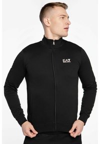 Giacca nera con zip e collo alto, maniche lunghe, tasche frontali e logo EA7 sul petto sinistro; realizzata in tessuto liscio e elastico.