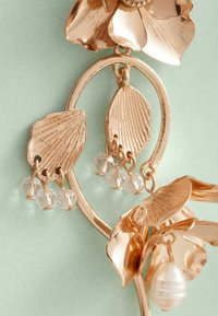 Design floreale in metallo rosato con foglie testurizzate, caratterizzato da perline trasparenti pendenti e un accento di perla. Sullo sfondo, un delicato verde.