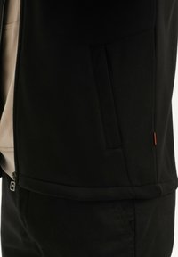 Primo piano di una persona che indossa una giacca nera con zip e pantaloni neri, evidenziando la tasca laterale della giacca e i dettagli della zip.