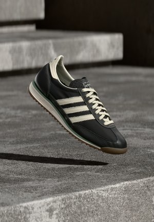 Must nahast toss, millel on kolm valget triipu, kreemjad paelad, tekstuuriga keel ja pruun kummist tallaosa. Minimalistlik disain retro mõjutustega.