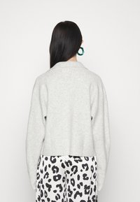Vrouw met lang, donker haar, gekleed in een lichtgrijze trui, zwart-witte luipaardprintbroek en groene oorringen, met haar rug naar de camera gekeerd.