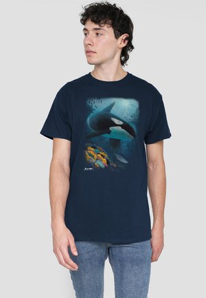 Marineblaues Baumwoll-T-Shirt mit einem grafischen Motiv eines Schwertwals, der über bunten Korallen und Fischen schwimmt, vor einem strukturierten ozeanischen Hintergrund.