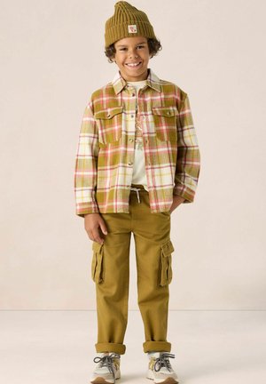 Chemise en flanelle à carreaux verts, roses et blancs ; t-shirt graphique crème ; pantalon cargo olive avec poches latérales ; bonnet en tricot jaune ; baskets grises.