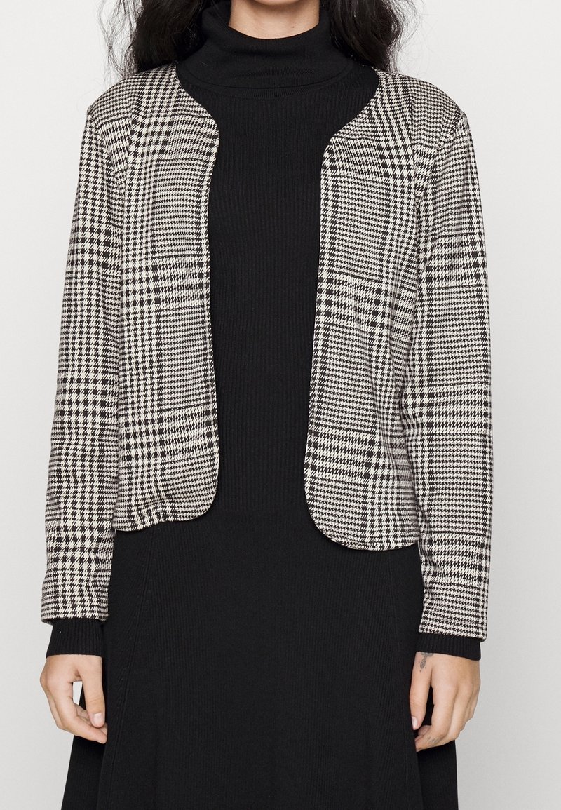 Femme portant une robe noire à col roulé et un blazer noir et blanc à motif pied-de-poule avec des manches longues.
