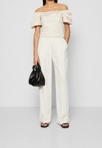 Top beige a spalle scoperte con maniche a sbuffo, abbinato a pantaloni bianchi a vita alta. Tiene una piccola borsa nera a texture.