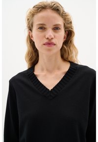 Maglione nero con scollatura a V e colletto a coste testurizzato. Design semplice, texture morbida e forma aderente. Nessun motivo o logo visibile.